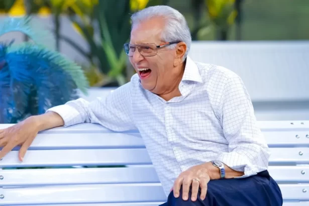 Carlos Alberto de Nóbrega, olhando para o esquerdo, dando um enorme sorrido, vestindo uma blusa branca e sentado no banco da praça