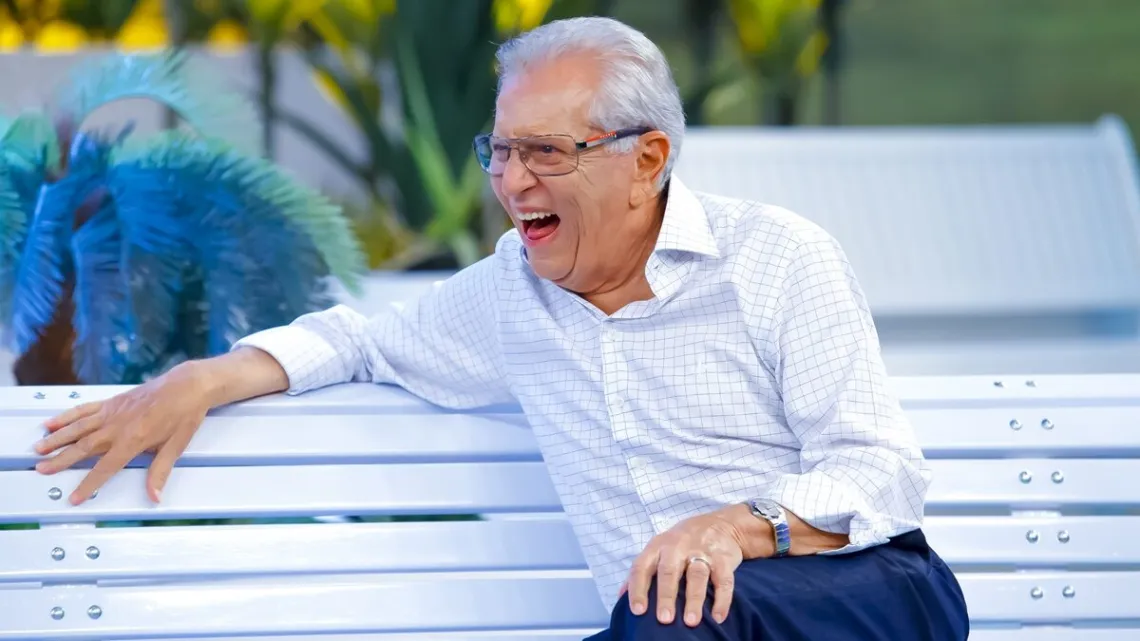Carlos Alberto de Nóbrega, olhando para o esquerdo, dando um enorme sorrido, vestindo uma blusa branca e sentado no banco da praça