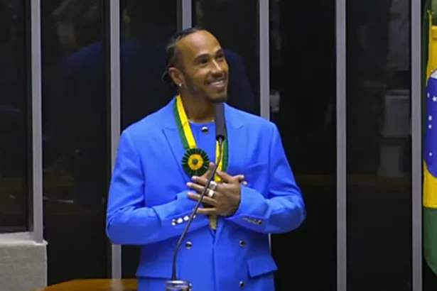 Lewis Hamilton, com um terno azul, de sorrido no rosto e com as mãos próximas ao peito, no plenário da câmara dos deputados