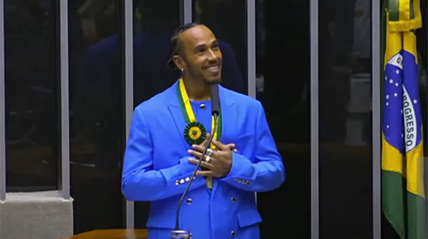 Lewis Hamilton, com um terno azul, de sorrido no rosto e com as mãos próximas ao peito, no plenário da câmara dos deputados