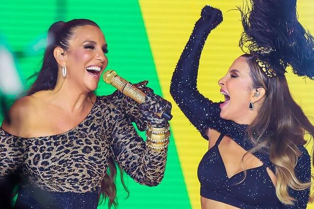 Claudia Leitte com o braço levantado, usando luva e top preto ao lado de Ivete Sangalo com look de onça e short preto segurando um microfone durante gravação de live de carnaval