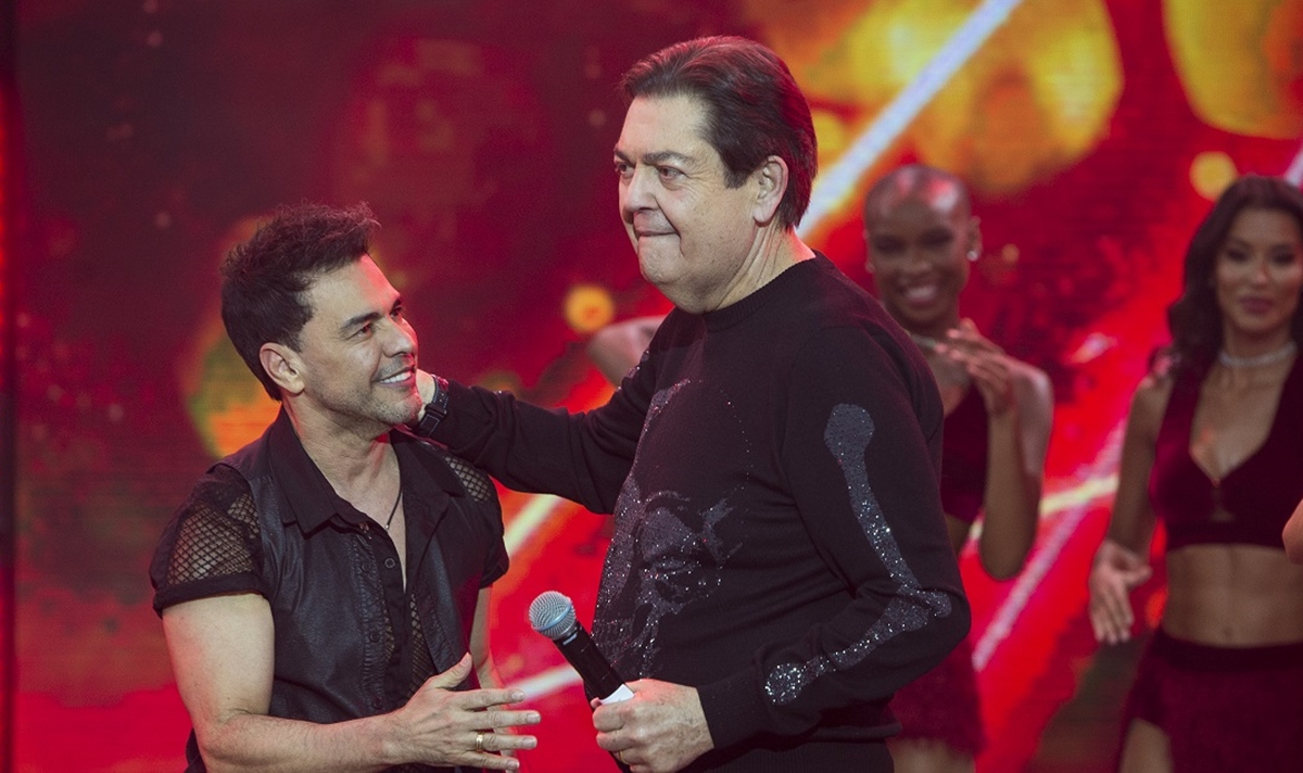 Zezé di Camargo usando camisa social preta, sorrindo, estendendo a mão esquerda pra Faustão; Faustão de camisa manga longa preta, segurando o micrfone com a mão esquerda e com a direta no pescoço de Zezé, durante gravação do Faustão na Band