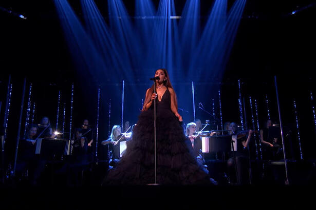 Ariana Grande vestida com vestido preto no palco de seu show, junto com orquestra feminina, em Londres.