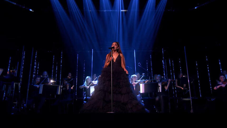 Ariana Grande vestida com vestido preto no palco de seu show, junto com orquestra feminina, em Londres.