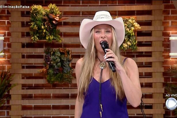 Adriane Galisteu de camiseta roxa, colar e chapéu de cowboy branco, segurando o microfone durante apresentação de A Fazenda 14, da Record TV