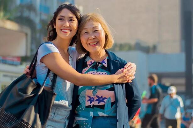 Ana Hikari sorrindo, com óculos de sol na cabeça, usando camisa branca e bolsa no ombro esquedo, abraçando a mãe durante viagem