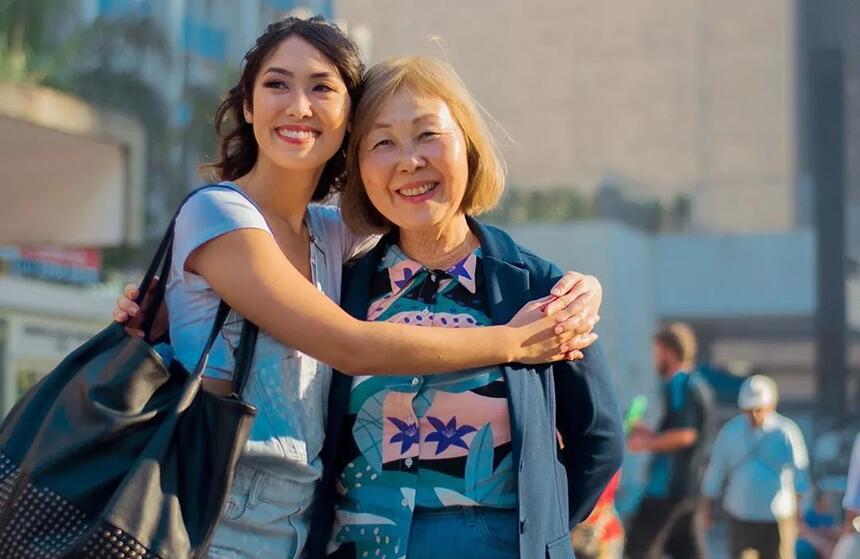 Ana Hikari sorrindo, com óculos de sol na cabeça, usando camisa branca e bolsa no ombro esquedo, abraçando a mãe durante viagem