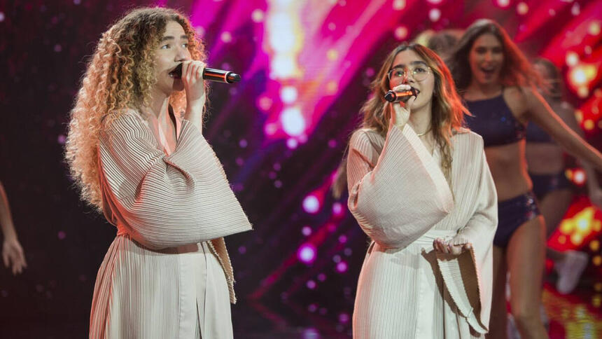 Anavitória com manta bege, segurando microfone durante performance no Faustão na Band