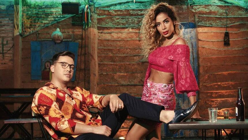 Wesley Safadão de camisa social estampada nas cores laranja, vermelho e branco, sentado, de óculos de grau, calça preta, com o pé esticado; Anitta com cabelo encaracolado, batom vermelho, saia rosa e tomara que caia em tom magenta, encostada numa mesa; ambos juntos para gravação de videoclipe