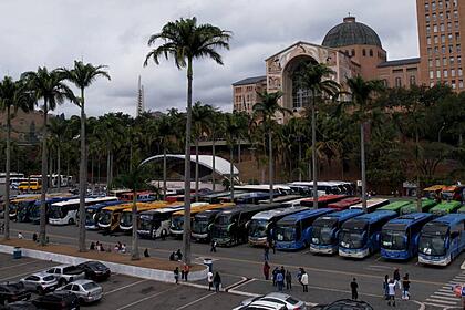 Estacionamento com carros, ônibus e pessoas. Ao fundo na cor marrom, está o Santuário Nacional de Nossa Senhora Aparecida, no interior de São Paulo.