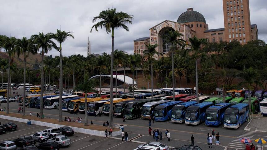 Estacionamento com carros, ônibus e pessoas. Ao fundo na cor marrom, está o Santuário Nacional de Nossa Senhora Aparecida, no interior de São Paulo.