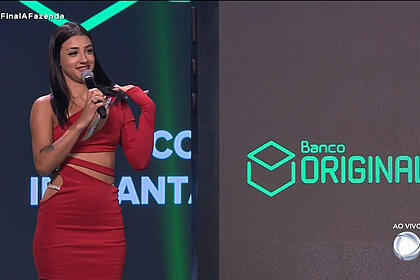 Bia Miranda com vestido vermelho, colar de microfone no pescoço, em pé, cabelo sollto, liso, batom vermeho, sentada durante gravação da final de A Fazenda 14, da Record TV