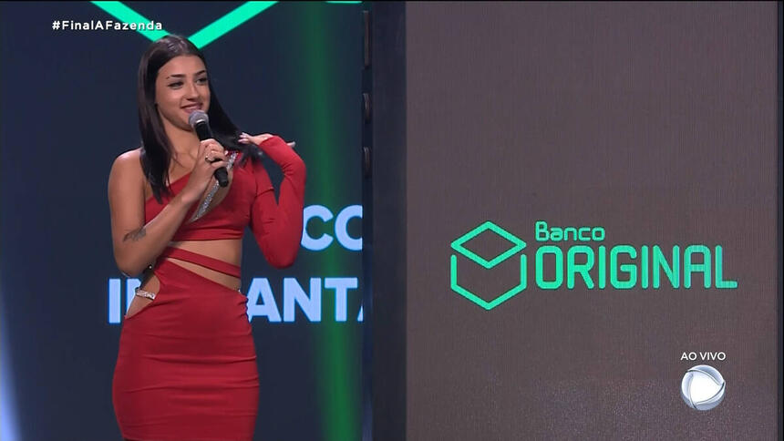 Bia Miranda com vestido vermelho, colar de microfone no pescoço, em pé, cabelo sollto, liso, batom vermeho, sentada durante gravação da final de A Fazenda 14, da Record TV