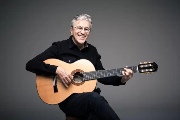 Caetano Veloso sorrindo, sentado, de camisa e calça preta, segurando um violão durante ensaio fotográfico