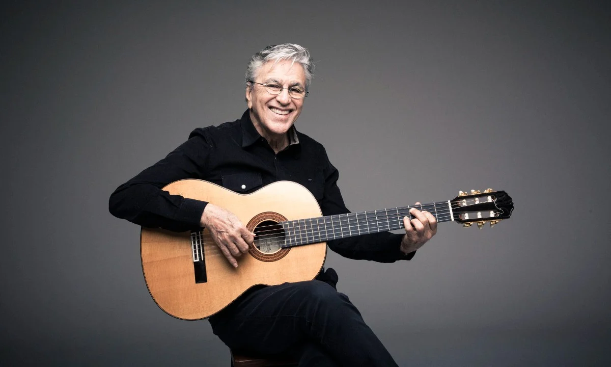 Caetano Veloso sorrindo, sentado, de camisa e calça preta, segurando um violão durante ensaio fotográfico