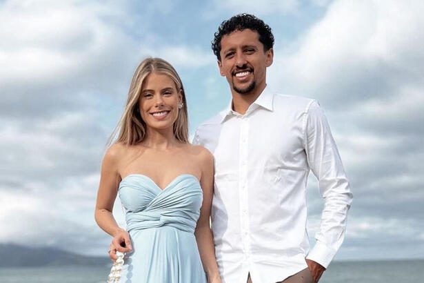 Carol Cabrino, com um vestido azul, ao lado de Marquinhos, com uma blusa social branca, em foto posada