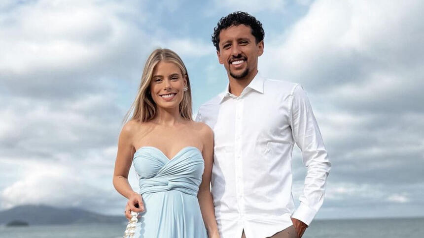 Carol Cabrino, com um vestido azul, ao lado de Marquinhos, com uma blusa social branca, em foto posada