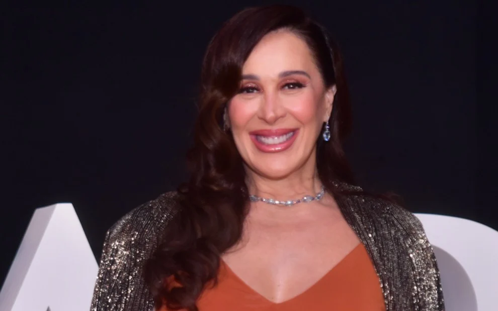 Claudia Raia com cardigã brilhoso, vestido marrom, sorrindo, com colar de brilhantes, cabelo solto durante desfile de uma marca de bijuterias