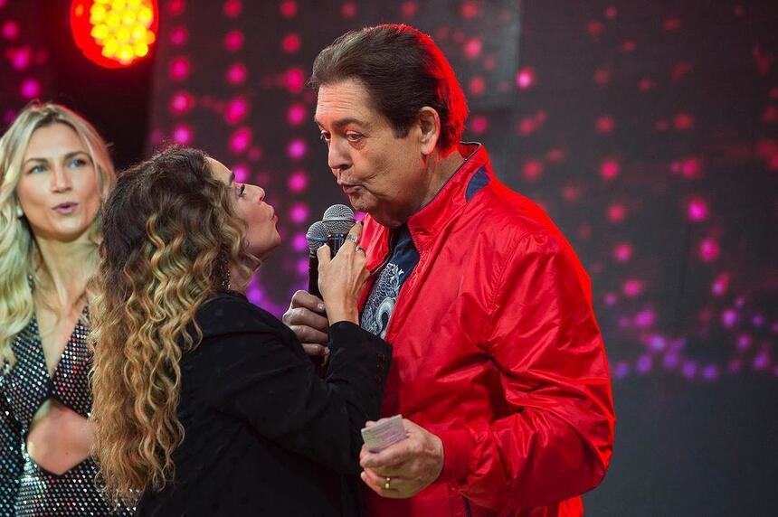 Daniela Mercury de blazer e top preto, puxando Faustão, de jaqueta vermelha, para dar um beijo, durante gravação de programa, na Band