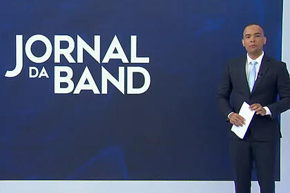 Diego Sarza, de terno, em pé em frente ao telão com o logo do Jornal da Band