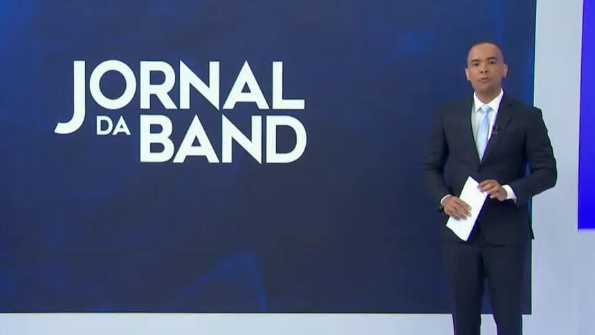 Diego Sarza, de terno, em pé em frente ao telão com o logo do Jornal da Band