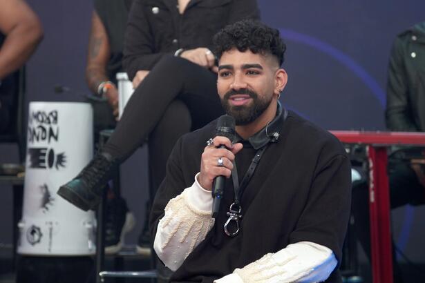 Dilsinho de camisa mangalonga preta, sorrindo, segurando o microfone durante entrevista no Altas Horas, da TV Globo