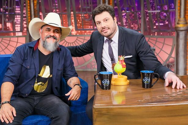 Donizeti Camargo de blazer azul marinho, camisa preta com estampa dourada, chapéu de cowboy branco, sorrindo, sentado num sofá azul ao lado de Danilo Gentili durante gravação do The Noite, do SBT