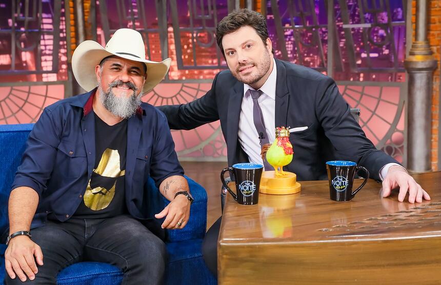 Donizeti Camargo de blazer azul marinho, camisa preta com estampa dourada, chapéu de cowboy branco, sorrindo, sentado num sofá azul ao lado de Danilo Gentili durante gravação do The Noite, do SBT
