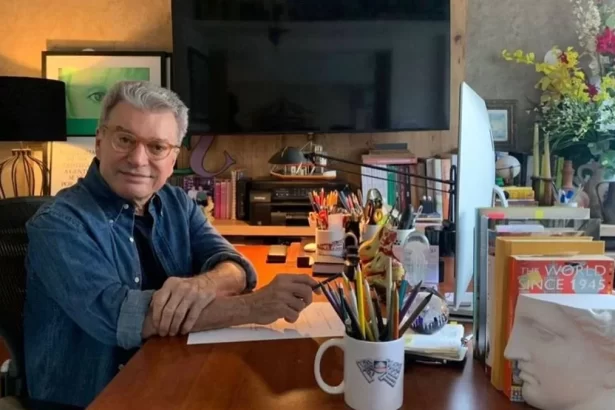 Edney Silvestre, sentado em frente uma mesa de escritório, com uma televisão ao fundo