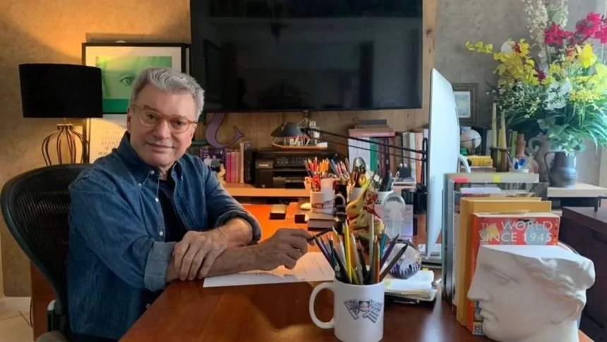 Edney Silvestre, sentado em frente uma mesa de escritório, com uma televisão ao fundo