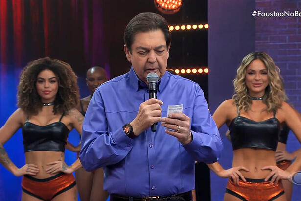 Faustão com uma blusa social azul, com bailarinas ao fundo, lendo um papel em uma das suas mãos, no estúdio do Faustão na Band