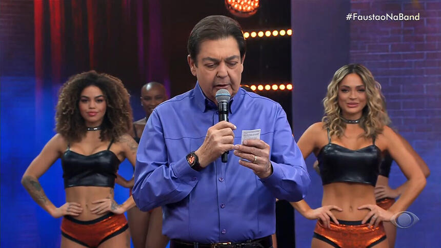 Faustão com uma blusa social azul, com bailarinas ao fundo, lendo um papel em uma das suas mãos, no estúdio do Faustão na Band