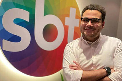 Fernando Jordão em foto posada, de braços cruzado, em frente ao logo do SBT