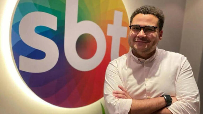 Fernando Jordão em foto posada, de braços cruzado, em frente ao logo do SBT