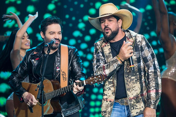 Fernando de jaqueta de couro, camisa preta, calça jeans, segurando violão; e Sorocaba de jaqueta estampada, chapéu de cowboy, calça jeans, segurando um microfone durante apresentação no palco do Faustão na Band