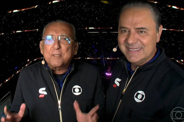 Galvão Bueno e Luís Roberto na cabine de transmissão da TV Globo no estádio Lusail, palco da final da Copa no Catar. Eles usam o uniforme de esporte da Globo e o estádio está com as luzes apagadas