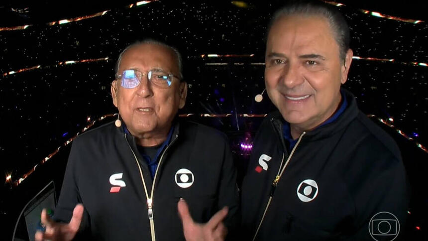 Galvão Bueno e Luís Roberto na cabine de transmissão da TV Globo no estádio Lusail, palco da final da Copa no Catar. Eles usam o uniforme de esporte da Globo e o estádio está com as luzes apagadas