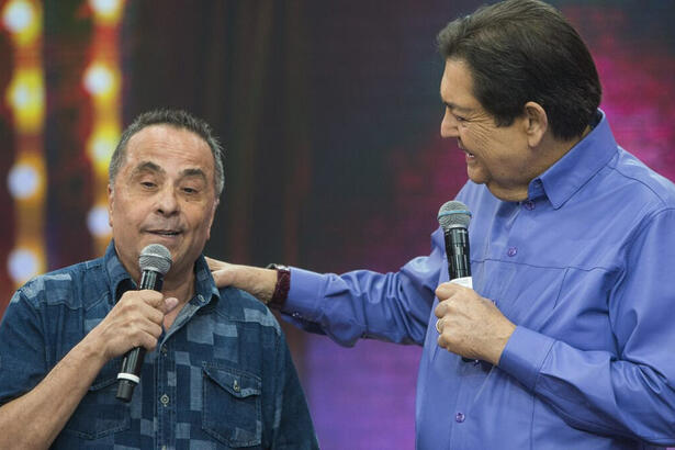 Geraldo Magela de camisa azul e preto estampada, segurando o micrfone ao lado de Fausto Silva, de camisa social azul, segurando no ombro de Geraldo, durante gravação do Faustão na Band