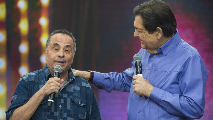 Geraldo Magela de camisa azul e preto estampada, segurando o micrfone ao lado de Fausto Silva, de camisa social azul, segurando no ombro de Geraldo, durante gravação do Faustão na Band