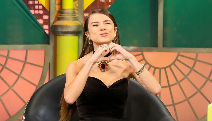 Giulia Be de top preto, colar, sentada num banco branco, fazendo coração com as mãos durante gravação de entrevista no The Noite, do SBT