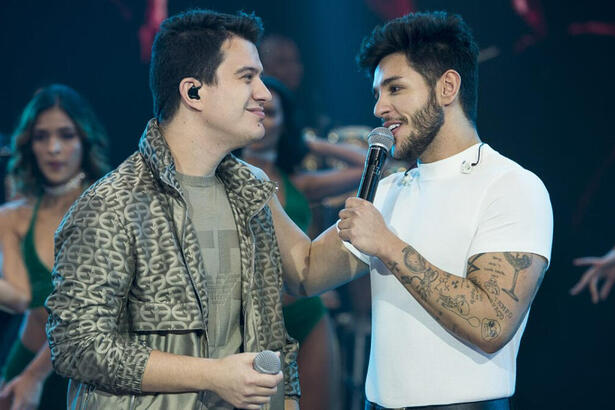 Hugo de jaqueta e camisa cinza segurando um microfone e Guilherme de camisa básica branca, segurando o microfone com a mão no ombro de Hugo durante performance no Faustão na Band