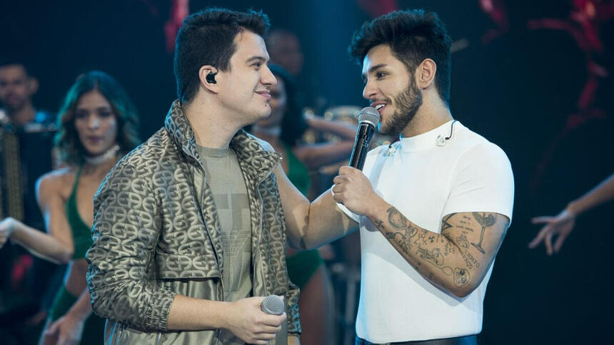 Hugo de jaqueta e camisa cinza segurando um microfone e Guilherme de camisa básica branca, segurando o microfone com a mão no ombro de Hugo durante performance no Faustão na Band