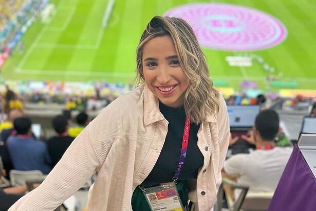 Isabelle Costa de jaqueta bege camisa preta e crachá, de cabelo solto, sorrindo, no estádio