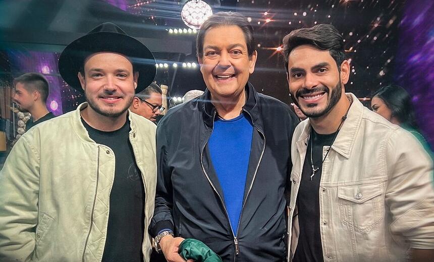 Israel de jaqueta bege e camisa preta sorrindo, de chapéu preto, ao lado de Faustão, de jaqueta preta e camisa azul, sorrindo, ao lado de Rodolffo, sorrindo, de jaqueta bege e camisa preta durante gravação do Faustão na Band