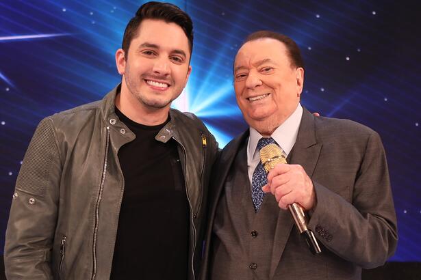 Jonas Esticado com jaqueta de couro preta, camisa básica preta, sorrindo, ao lado de Raul Gil durante gravação de programa no SBT