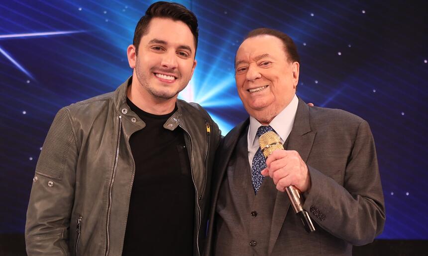 Jonas Esticado com jaqueta de couro preta, camisa básica preta, sorrindo, ao lado de Raul Gil durante gravação de programa no SBT