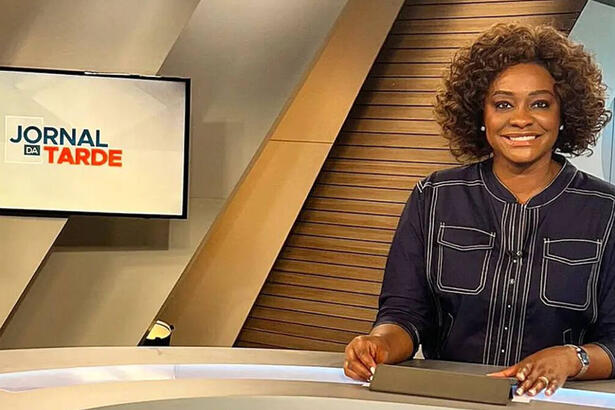 Joyce Ribeiro com uma blusa escura, sentada na bancada do Jornal da Tarde, na TV Cultura, com televisor ao fundo