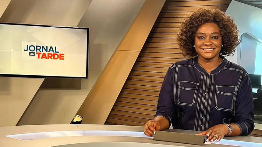 Joyce Ribeiro com uma blusa escura, sentada na bancada do Jornal da Tarde, na TV Cultura, com televisor ao fundo