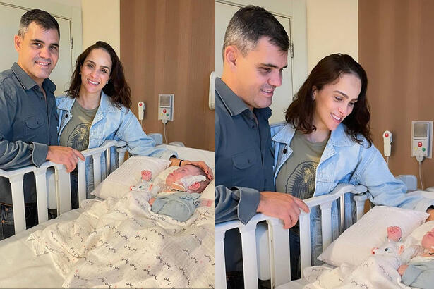 Juliano Cazarré com jaqueta jeans azul sorrindo, ao lado da esposa de camisa verde musgo e jaqueta azul jeans, sorrindo, com a filha deitada numa cama pós-cirurgia