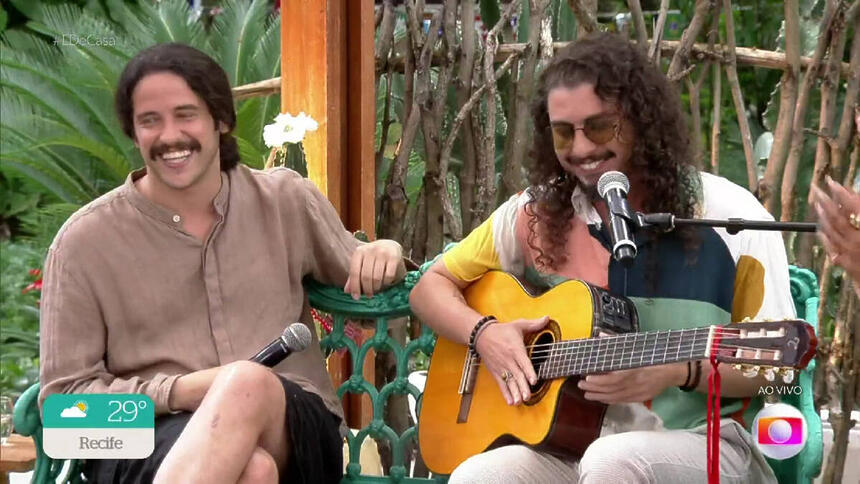 Juzé de óculos de sol, cabelo solto, segurando o violão; Lukete sorrindo, com cabelo preso, perna cruzada duraante gravação do É de Casa